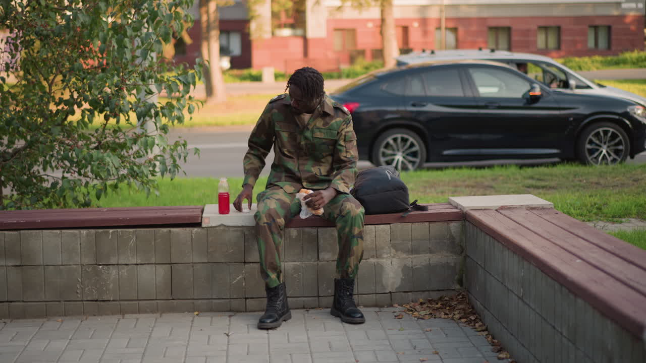 Black Soldier In Camouflage Eating Sandwich On Concrete Ledge With Red Drink At Side, Adjusting Boots And Jacket, Late Afternoon Light, Passing Traffic And Suburban Buildings, Quiet Relief