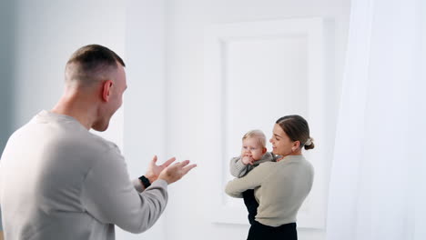 happy woman holding a baby in hands. man claps his hands to attract child's attention. father reaches his arms to a son and kid grabs dad's finger.