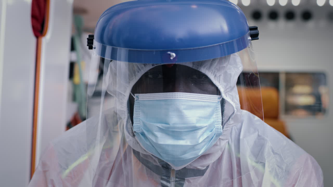 african American Paramedic in Hazmat Suit Looking at Camera Weary Black Male Medical Practitioner in Hazmat Suit with Mask and Face Shield Looking at Camera while Riding Ambulance to Emergency during Pandemic
