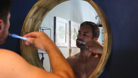 guy in 30s brushing teeth in bathroom, over shoulder view in round mirror