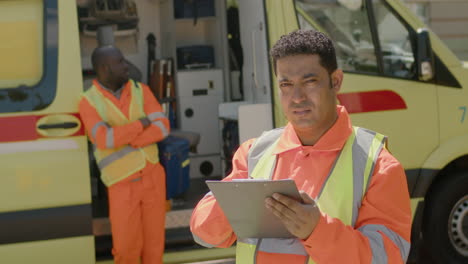 Latin-Medical-Assistance-Writing-On-A-Clipboard-While-His-Colleague-Is-On-The-Ambulance-With-Arms-Crossed