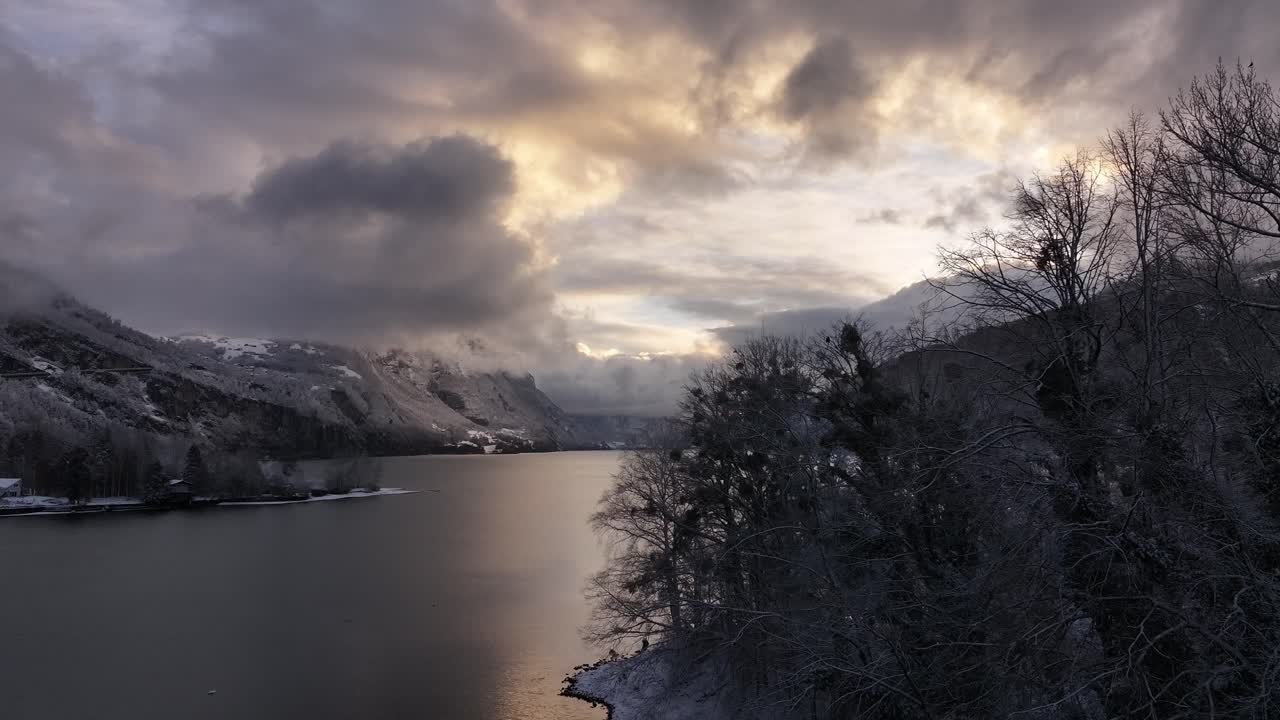 A tranquil late evening aerial view of Switzerland's Walensee Lake, surrounded by snow-dusted trees and mountains under dramatic, cloudy skies, highlighting serene and moody winter beauty.