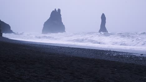 Weite-Aufnahme-Der-Berühmten-Felsformationen-Des-Schwarzen-Sandstrandes-Von-Reynisfjara-An-Einem-Bewölkten-Winterabend-Mit-Riesigen-Wellen,-Die-Zusammenbrechen