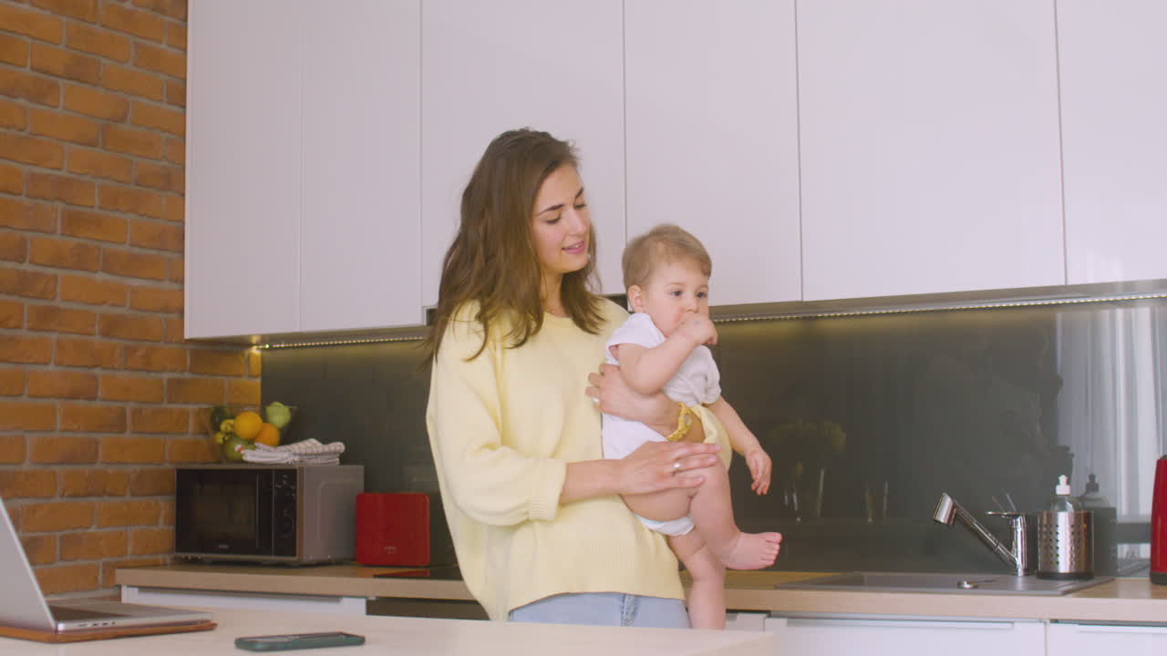 Smiling Woman Holding Her Baby In The Kitchen, Then She Sits In Front Of The Laptop