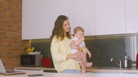 Smiling-Woman-Holding-Her-Baby-In-The-Kitchen,-Then-She-Sits-In-Front-Of-The-Laptop