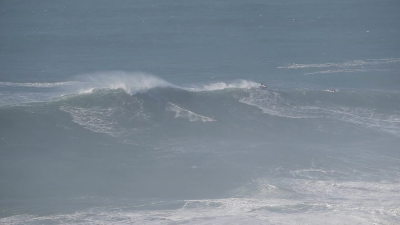 Enormous wave crashing on the shore in Nazare, Portugal, surfers watching and one of them riding it.