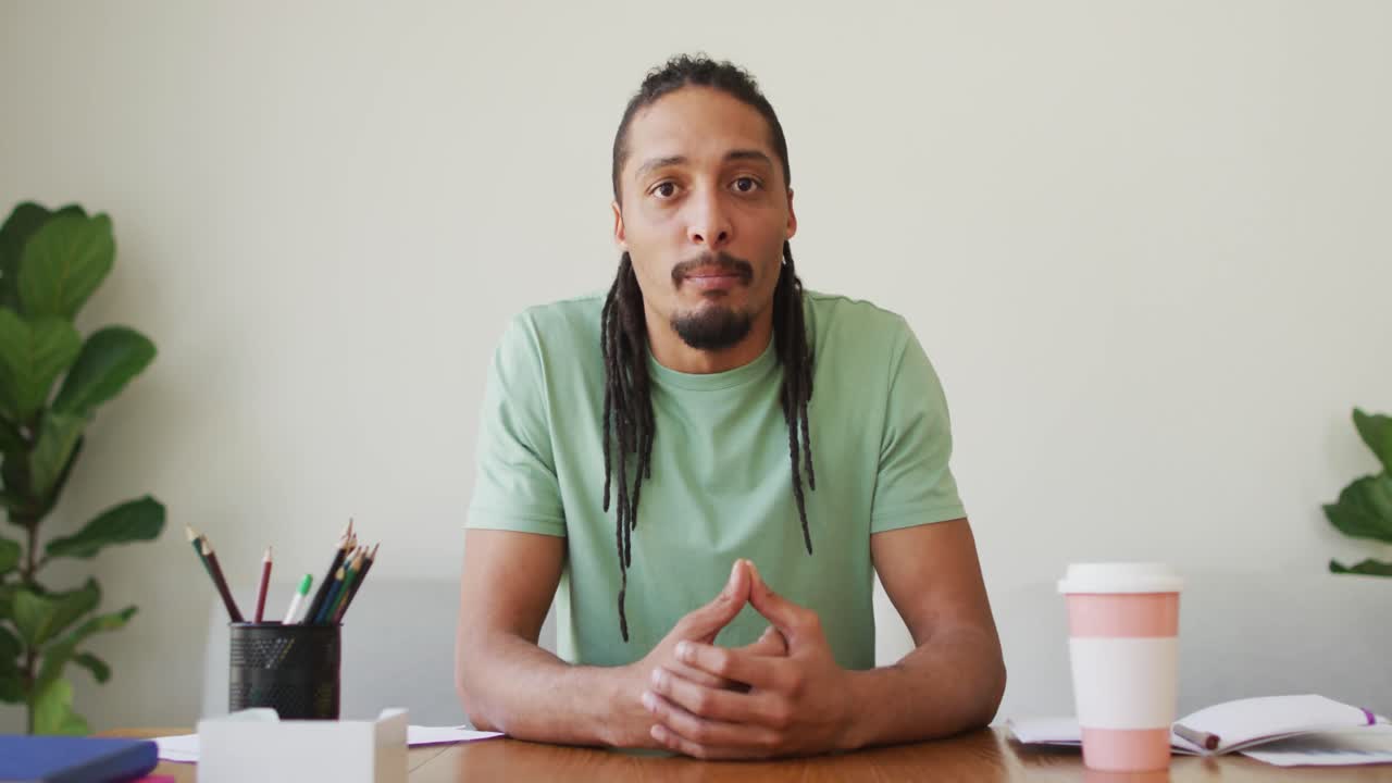 Happy biracial man with dreadlocks sitting at desk, smiling and waving during video call