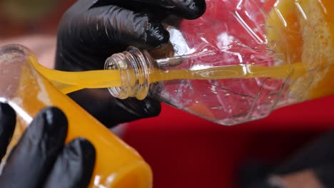 person in black gloves pouring orange juice from a plastic bottle into a glass bottle