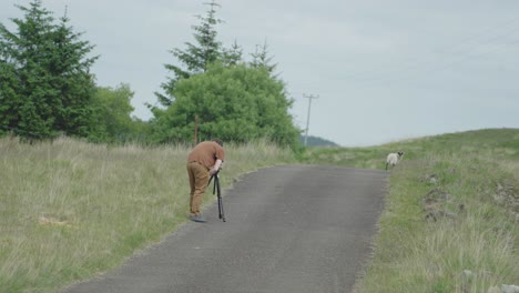 man using a camera in a tripod to capture footage of sheep at roadside