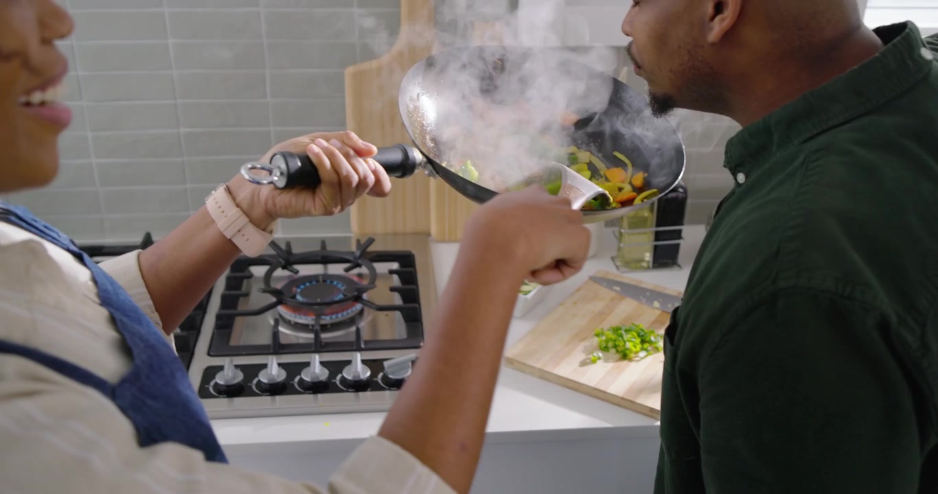 Couple afro-américain dans une cuisine, cuisinant au wok et offrant des légumes à son partenaire qui arrive pour partager