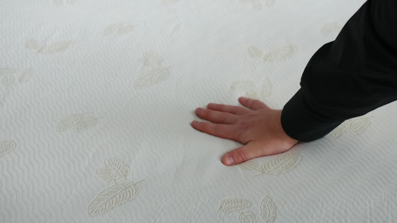 A hand pressing firmly on a white mattress with embossed leaf patterns and a soft wavy texture. Close-up capturing fabric compression and mattress responsiveness under pressure
