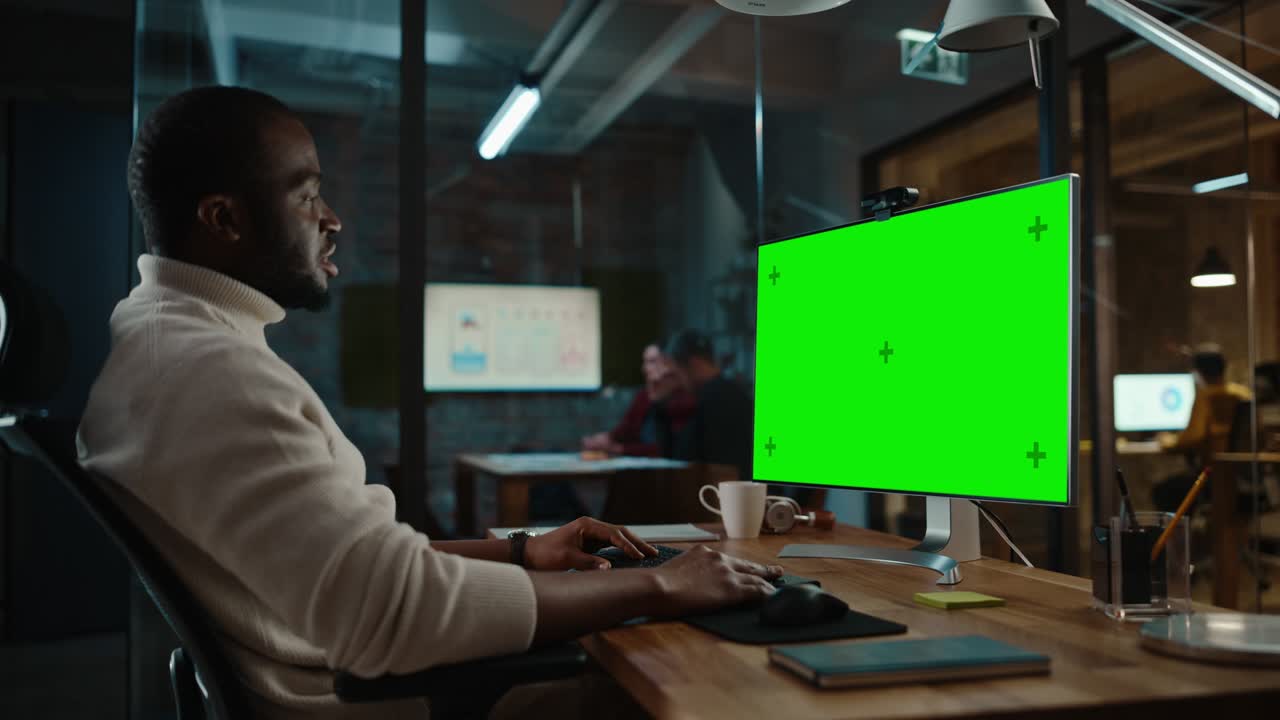 el guapo gerente de proyecto afroamericano está haciendo una llamada de video en una computadora de escritorio con pantalla verde en una oficina creativa ocupada.