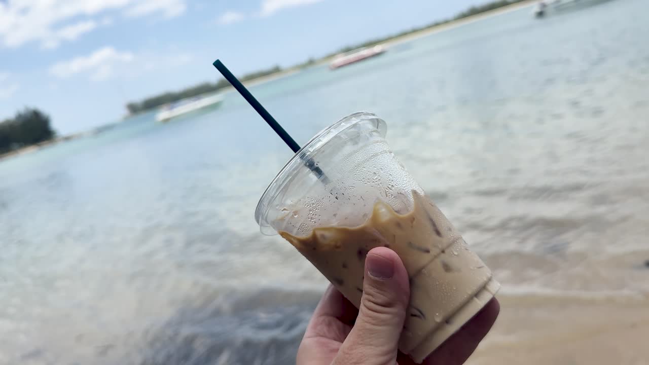 Hand holding iced coffee cup by calm ocean on sandy beach under bright daylight
