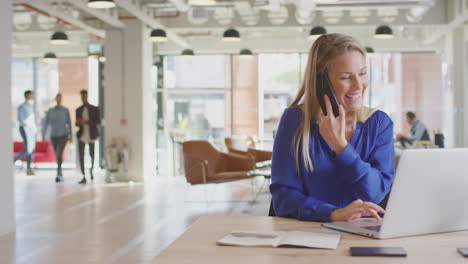 businesswoman sitting at desk on phone call in modern open plan office with colleagues in background