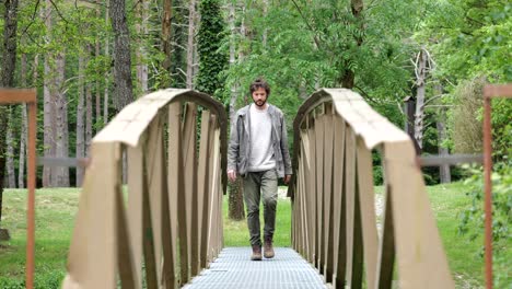 Young-bearded-man-in-his-20s-walking-over-a-small-footbridge-towards-the-camera