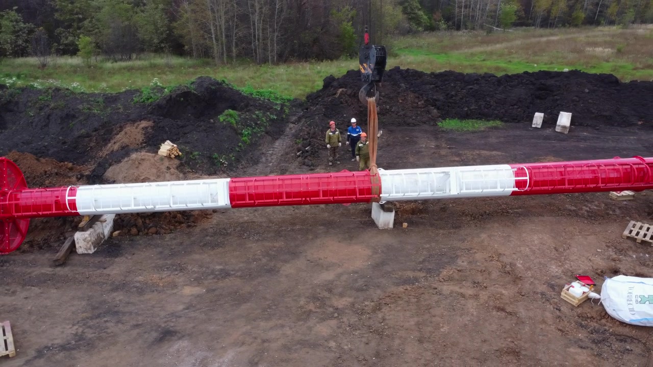 Grande sezione di condotta rossa e bianca giace in un cantiere fangoso con operai nelle vicinanze e un gancio di gru sospeso in alto, circondato da terreno scuro