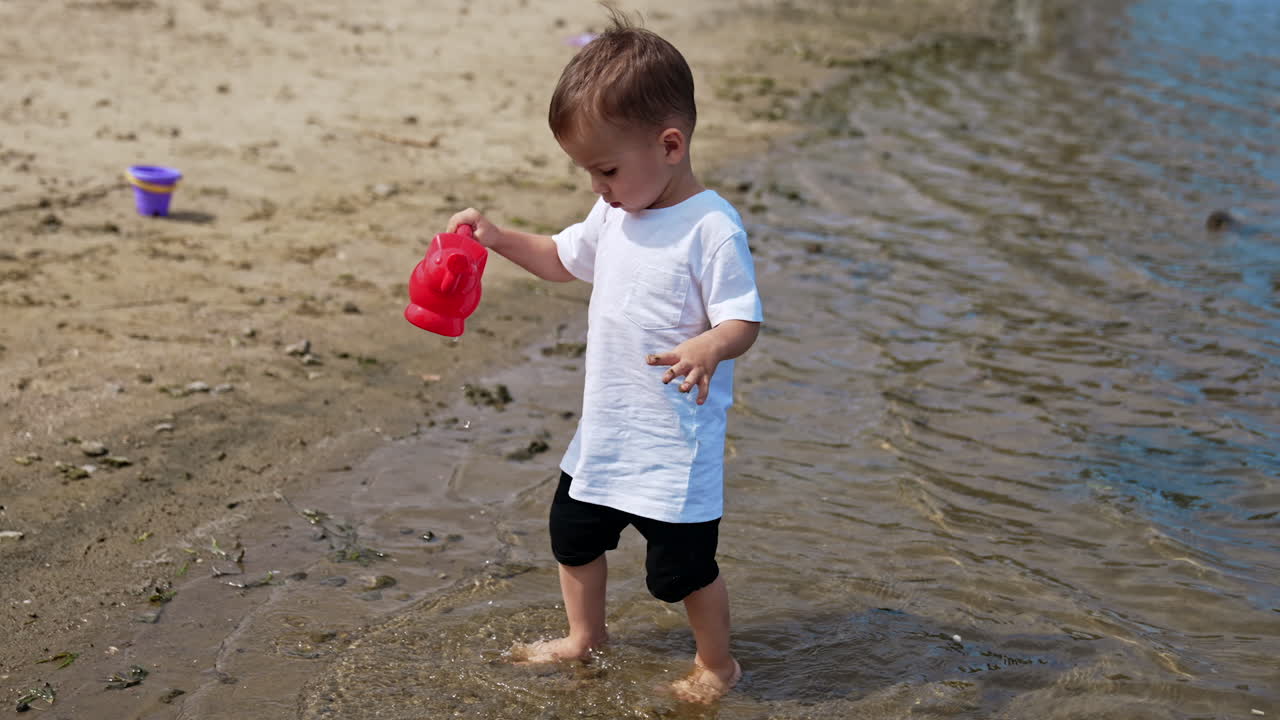 Cute toddler picks up some water with his red watering can. Little baby carries funnel carefully to his bucket on the beach.