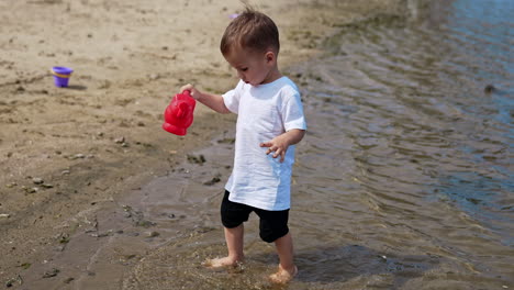 cute toddler picks up some water with his red watering can. little baby carries funnel carefully to his bucket on the beach.