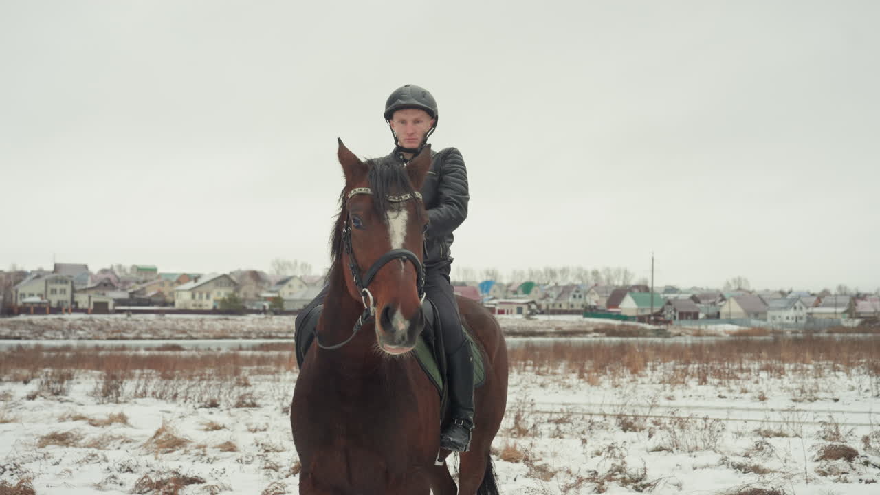 Custode caucasico che conduce e calma il cavallo prima di montare in un prato innevato. Mano delicata sulla briglia, risposta paziente del cavallo, sfondo rurale panoramico con l'orizzonte di un villaggio, legame emotivo e quiete.
