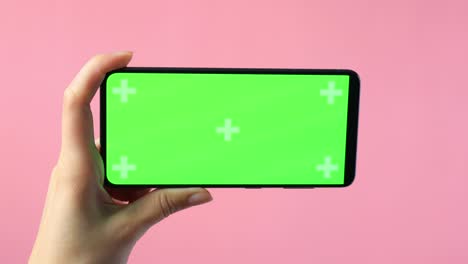 close-up of female hands holding smartphone with blank green screen showing copy space for app or website design, on pink background , mockup.