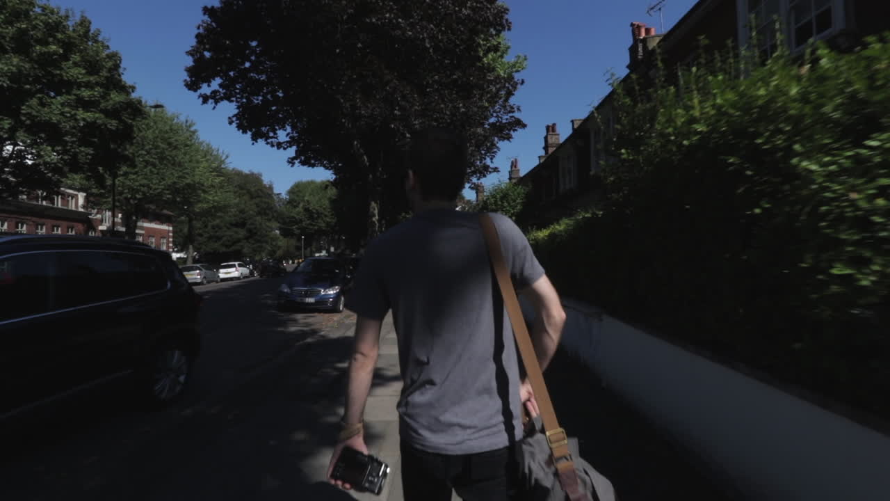 Kameraføring der følger en kaukasisk mand, iført grå T-shirt med kamerataske slynget over den ene skulder og bærende et kamera, mens han går ned ad en gade i Highgate, Nordlondon, Storbritannien, på en solrig dag.