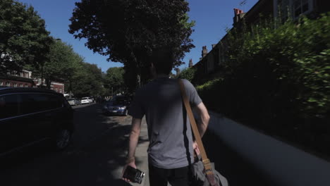 moving shot following caucasian man, wearing grey t shirt with camera bag slung over one shoulder carrying camera, walking down street in north london highgate uk day time sunny