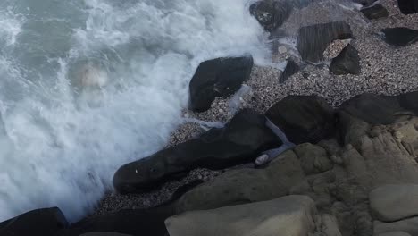 Waves-splashing-and-foaming-on-black-carved-rocks-plunged-in-beach-with-pebbles,-dramatic-seascape-in-coastline-of-Ionian-sea-in-Albania