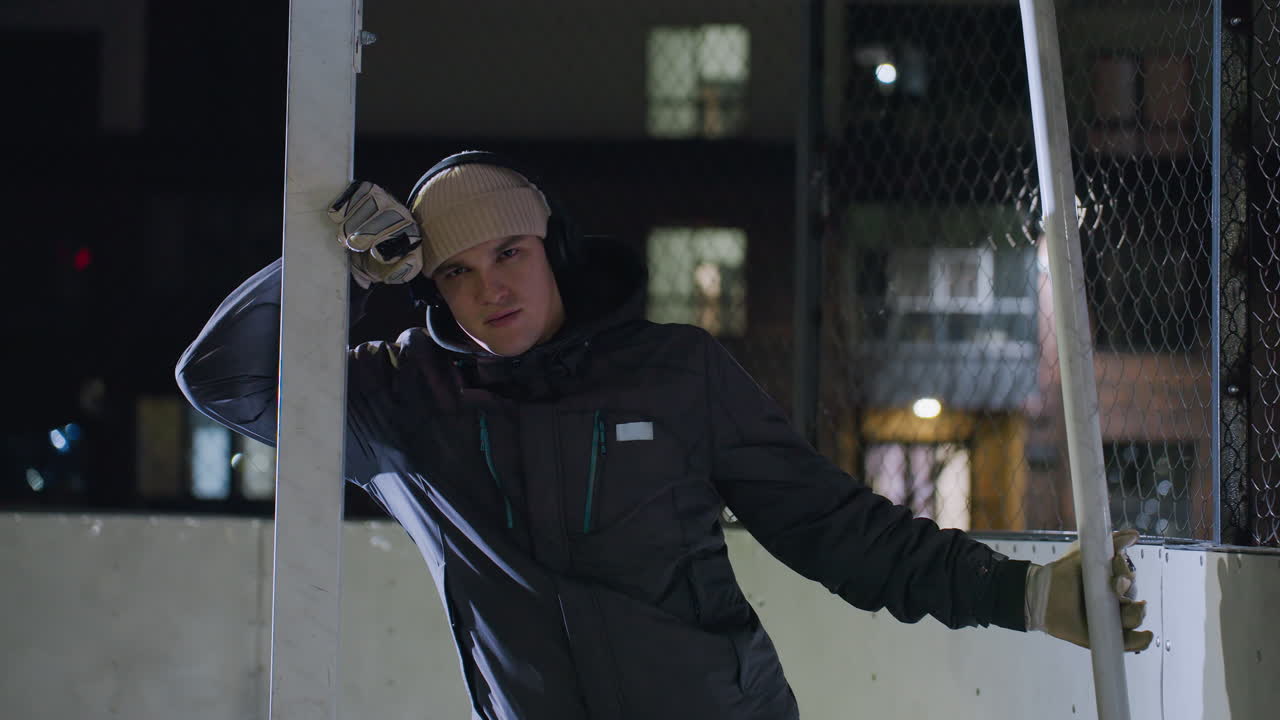 골 포스트에 기대어, 장갑, 베니, 그리고 헤드폰을 입은 피곤한 남자가 밝은 밤 조명 아래, 체인 링크 울타리 배경에서 반사되는 도시 조명에 집중하고 있습니다.