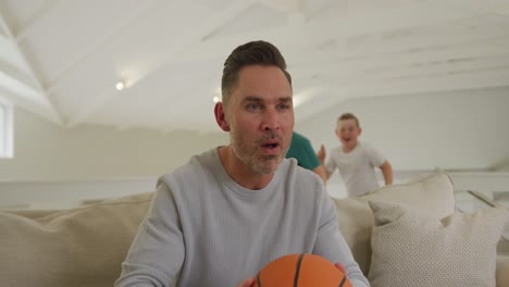 two caucasian boys hugging their father holding basketball and sitting on the couch at home
