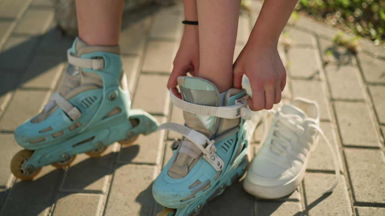Close-up of someone putting left leg into cyan roller skate, adjusting foot for secure fit, white sneaker placed nearby on pavement under warm sunlight, with green natural background slightly blurred