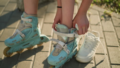 close-up of someone putting left leg into cyan roller skate, adjusting foot for secure fit, white sneaker placed nearby on pavement under warm sunlight, with green natural background slightly blurred