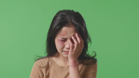 close up of young asian woman victim of violence with bruise on face covering her hurt eye and crying on green screen background in the studio