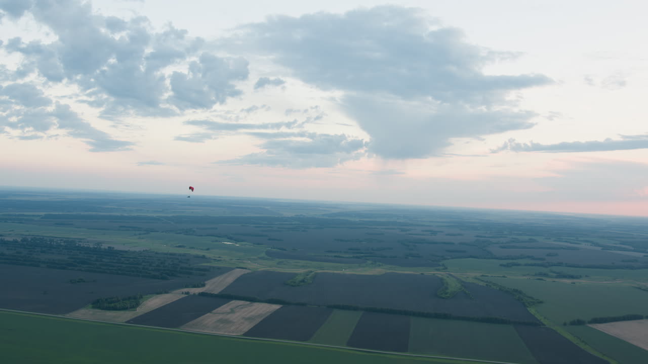 Distant silhouette of pilot flying under paraglider canopy drifting across pastel sky at sunset, clouds framing peaceful aerial drift above patchwork fields and tree lines in evening light