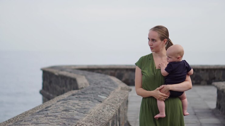 A young mother and her baby son stroll along the waterfront near the ocean on an ancient European square, enjoying the view of the waves and smiling joyfully