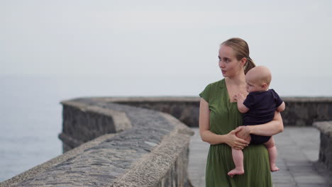 A-young-mother-and-her-baby-son-stroll-along-the-waterfront-near-the-ocean-on-an-ancient-European-square,-enjoying-the-view-of-the-waves-and-smiling-joyfully