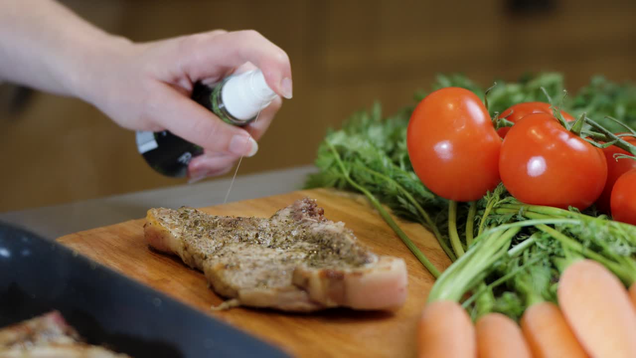 Hand sprays marinade over raw spiced meat with vegetables on kitchen counter