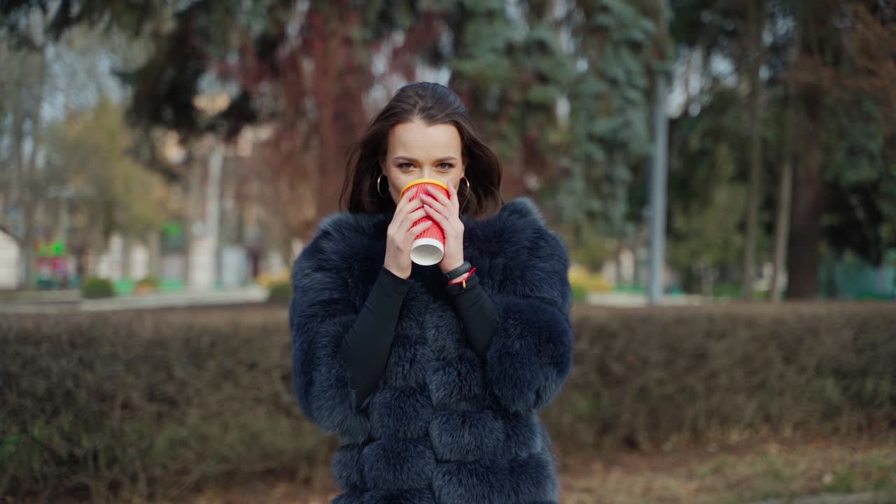 Beautiful girl drinks coffee outdoors. Portrait of a fashionable woman in fur coat with hot drink looking on camera. Stylish model in the park.