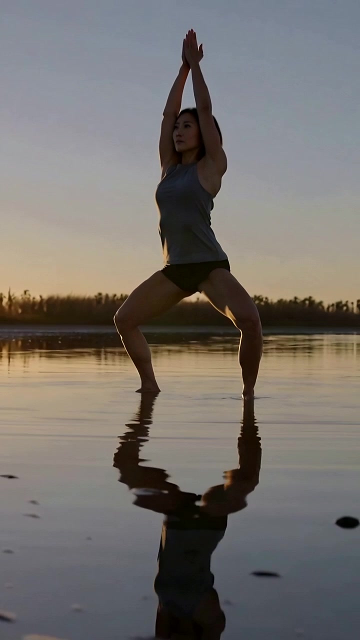 Video verticale: Donna in canottiera e pantaloncini che si bilancia e alza le braccia sulla spiaggia al tramonto, riflesso