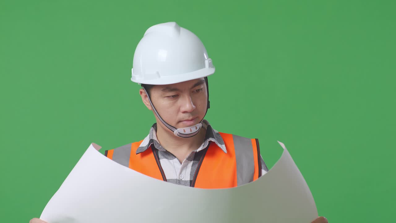 Close Up Of Asian Male Engineer With Safety Helmet Looking At Blueprint In His Hands And Looking Around While Standing In The Green Screen Background Studio