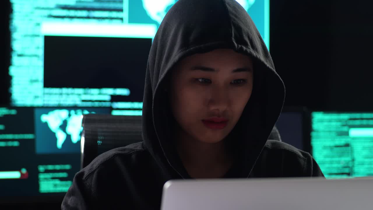 primo piano di una hacker donna asiatica con un cappuccio che guarda la telecamera e incrocia le braccia mentre lavora su un portatile con mappe e dati su schermi di più monitor in una stanza d'ufficio buia