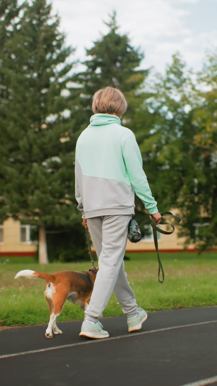 Persona paseando a un beagle por un sendero, vista trasera de un adulto con sudadera sujetando la correa, paso relajado junto al perro, árboles y pavimento del vecindario, paseo de bienestar y vínculo de compañía, actividad casual al aire libre