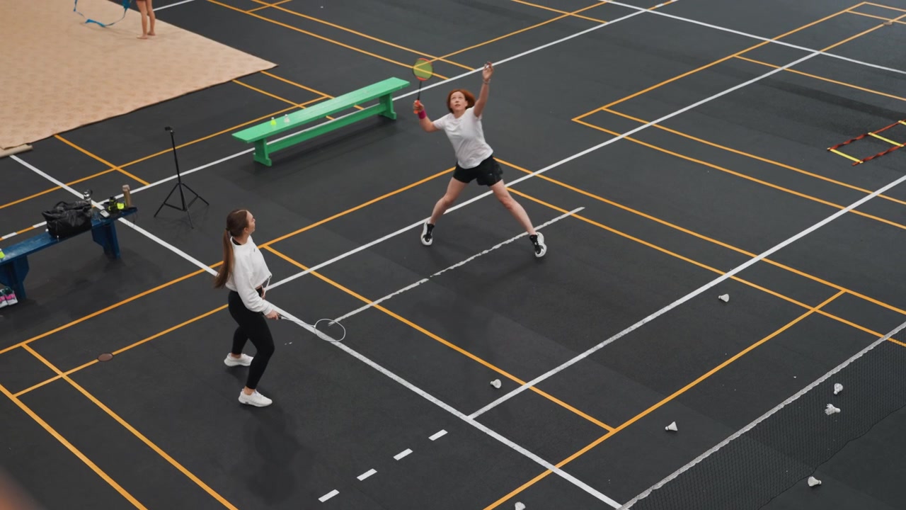 Due donne bianche su un campo da badminton impegnate in un rapido scambio durante una sessione di allenamento indoor, volano a mezz'aria, gioco di gambe concentrato e scambi a rete, panca verde e attrezzatura a bordo campo, energico.
