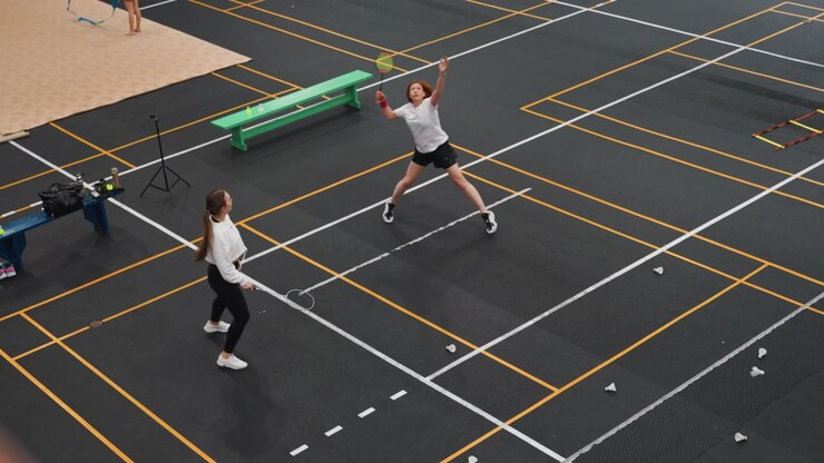 Due donne bianche su un campo da badminton impegnate in un rapido scambio durante una sessione di allenamento indoor, volano a mezz'aria, gioco di gambe concentrato e scambi a rete, panca verde e attrezzatura a bordo campo, energico.