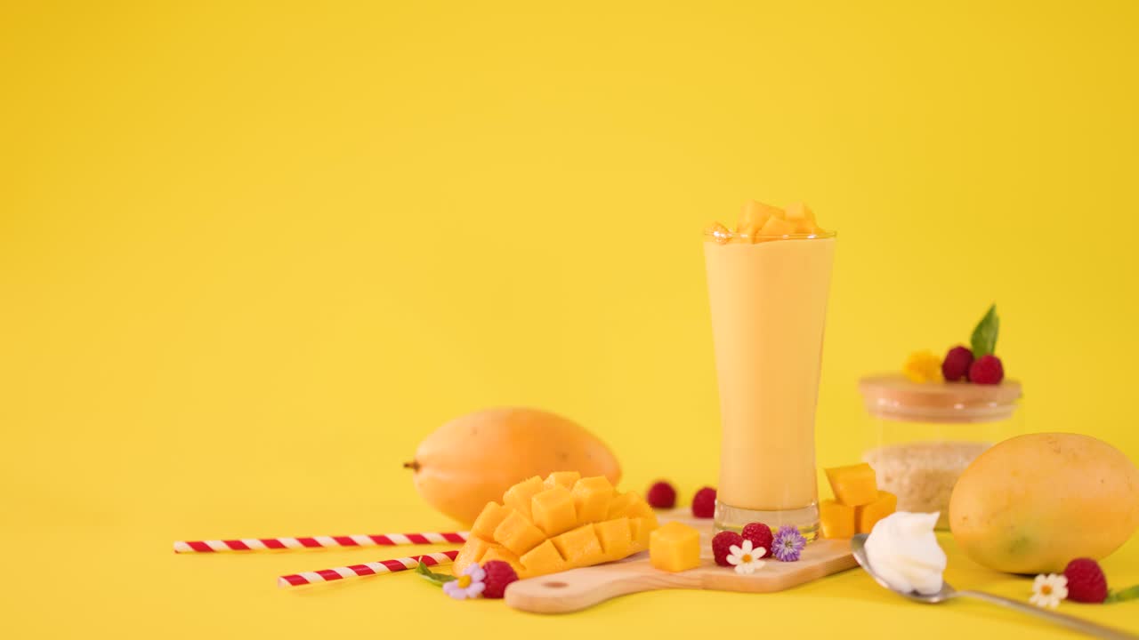 Human hand adds mint garnish to tall mango smoothie among tropical fruits, bright studio lighting