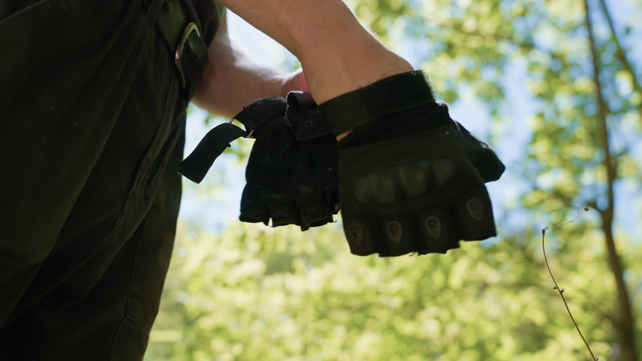 Under view of soldier wearing right hand fingerless tactical glove with flies perching on knuckle protector, sunlight filtering through green foliage and blue sky backdrop framing wrist detail
