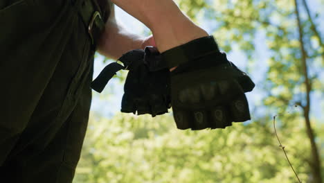 under view of soldier wearing right hand fingerless tactical glove with flies perching on knuckle protector, sunlight filtering through green foliage and blue sky backdrop framing wrist detail