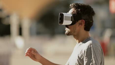 vista lateral de un joven árabe con cabello rizado oscuro y barba en camiseta gris estando en el parque con gafas de realidad virtual