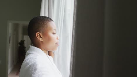 Happy-african-american-plus-size-woman-wearing-robe,-opening-curtains-and-looking-through-window