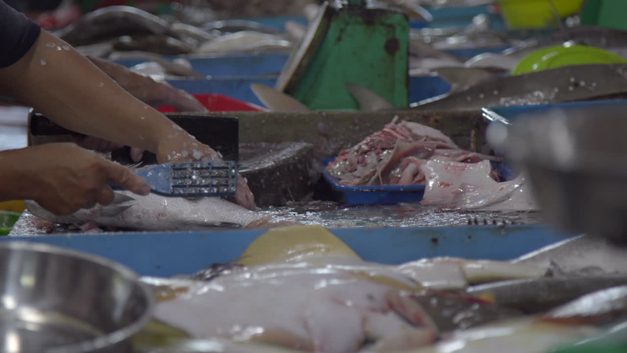 Close-up shot of a market vendor manually and rapidly descaling a fresh, raw fish on a busy, unsanitary counter. Illustrates the labor and preparation required in the Asian seafood supply chain