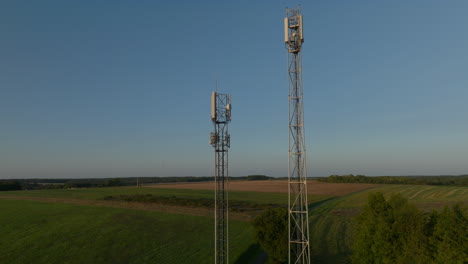 Two-radio-towers-in-the-middle-of-farmland-during-sunrise,-aerial-orbital-dolly-in-tilting-upward